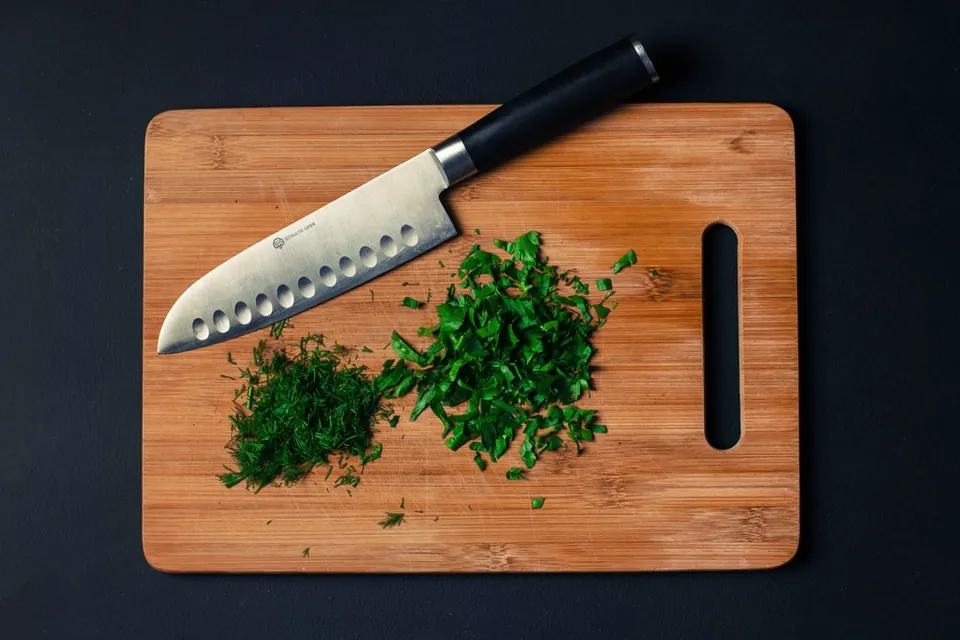 A santoku knife with a scalloped blade beside chopped herbs on a bamboo cutting board