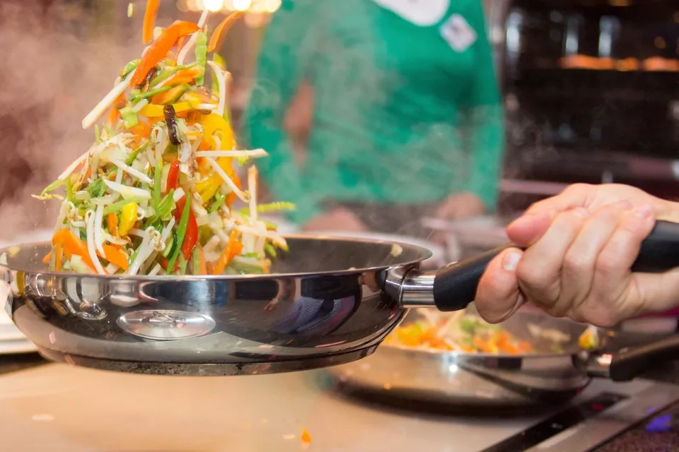 A cook tossing sliced vegetables in a stainless steel frying pan over a hob