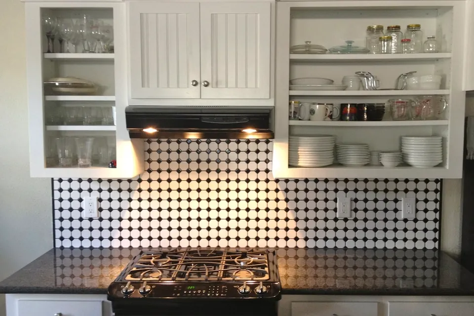 Open kitchen shelves of glasses, plates and jars above a gas hob and tiled splashback