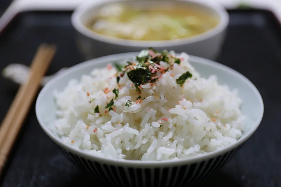 A bowl of steamed white rice topped with seaweed flakes, with chopsticks and a bowl of soup behind