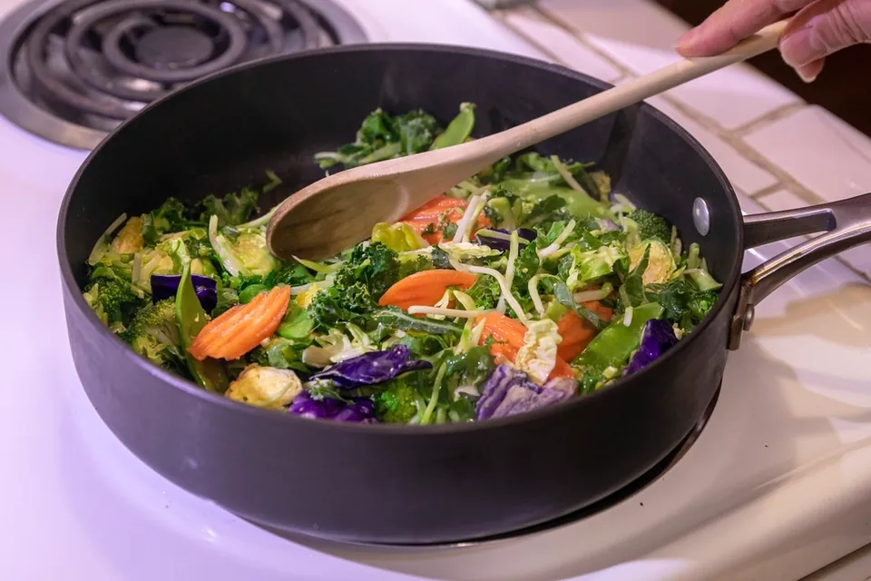 Vegetables being stirred with a wooden spoon in a dark nonstick saute pan on an electric hob