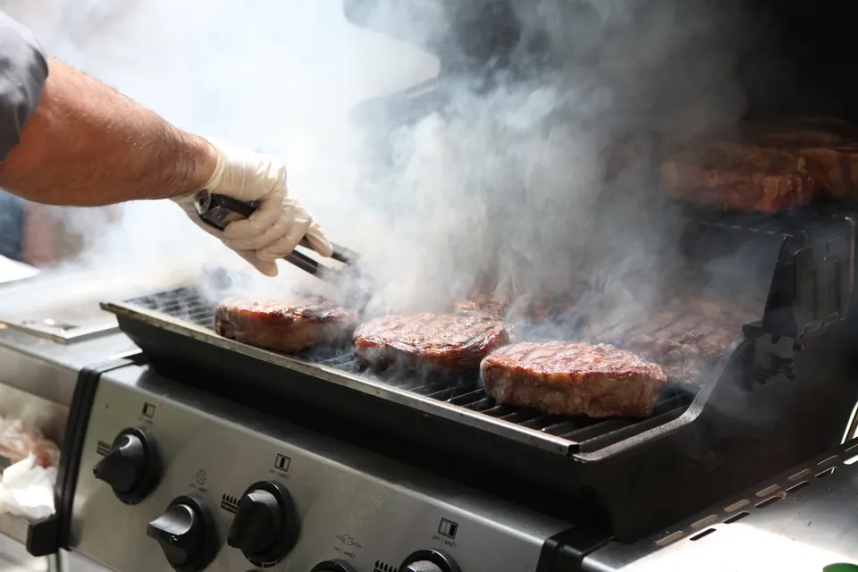 Burgers searing on a gas grill, a gloved hand holding tongs