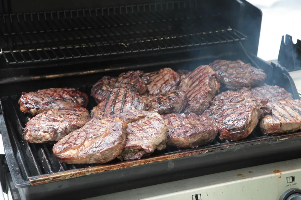 Steaks cooking on the grates of a gas grill with the lid open